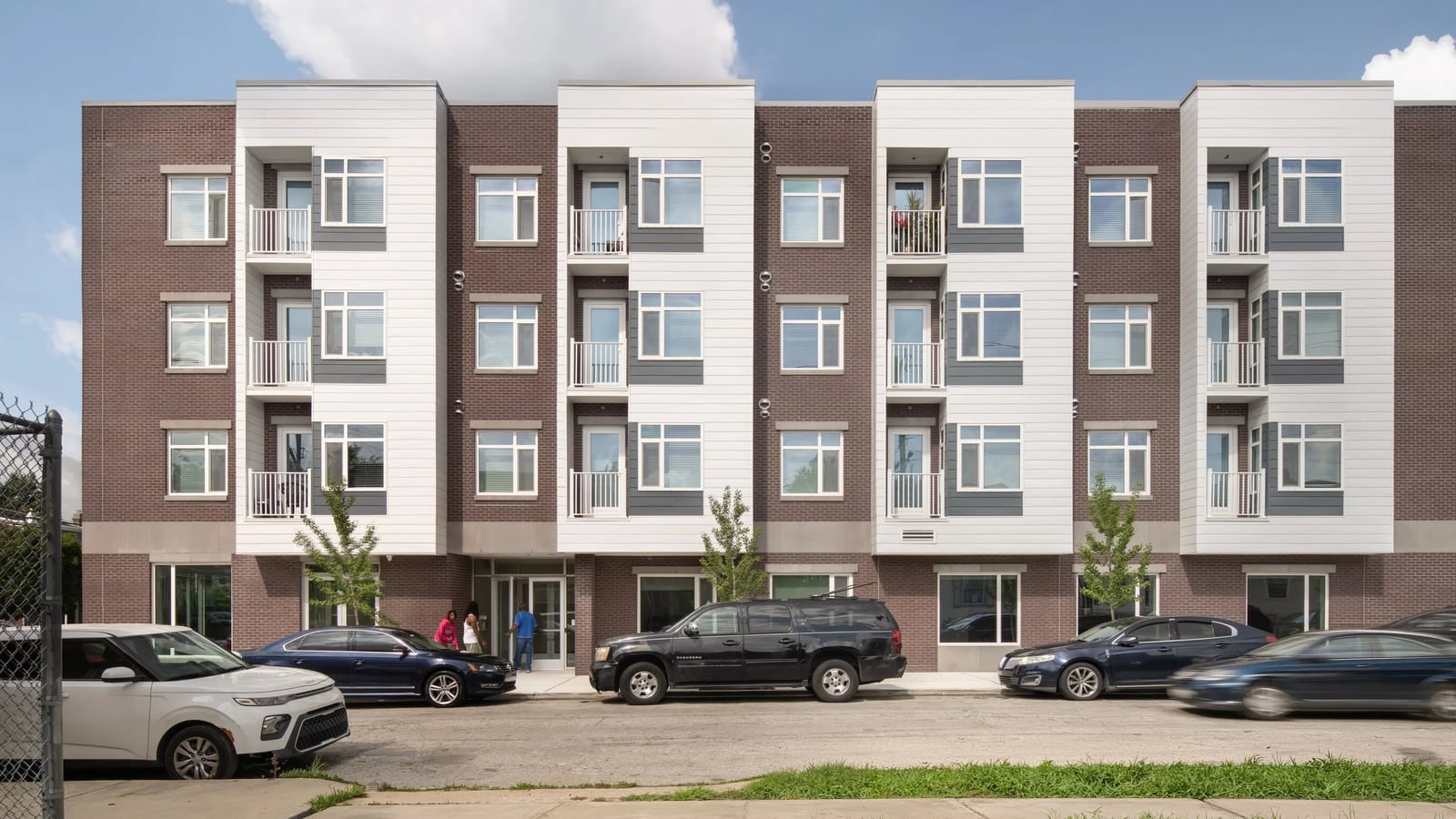 Long side of a four-story building in brown brick with four white bays of balconies, a glass entrance where three people stand, young street trees and cars parked at the curb