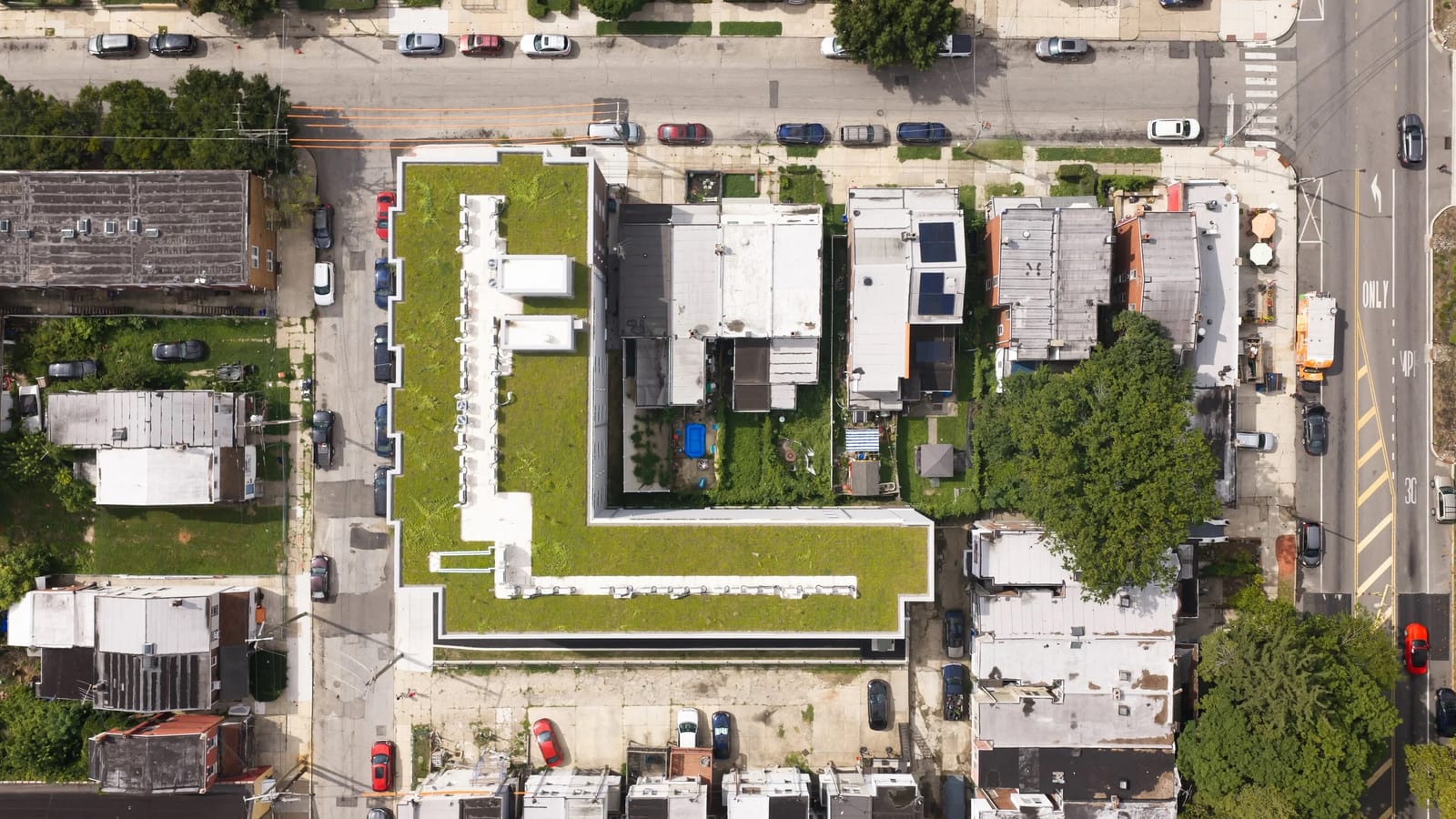 Overhead photograph of an L-shaped building with a planted green roof and a row of white rooftop equipment, with rowhouses, a parking area and streets around it