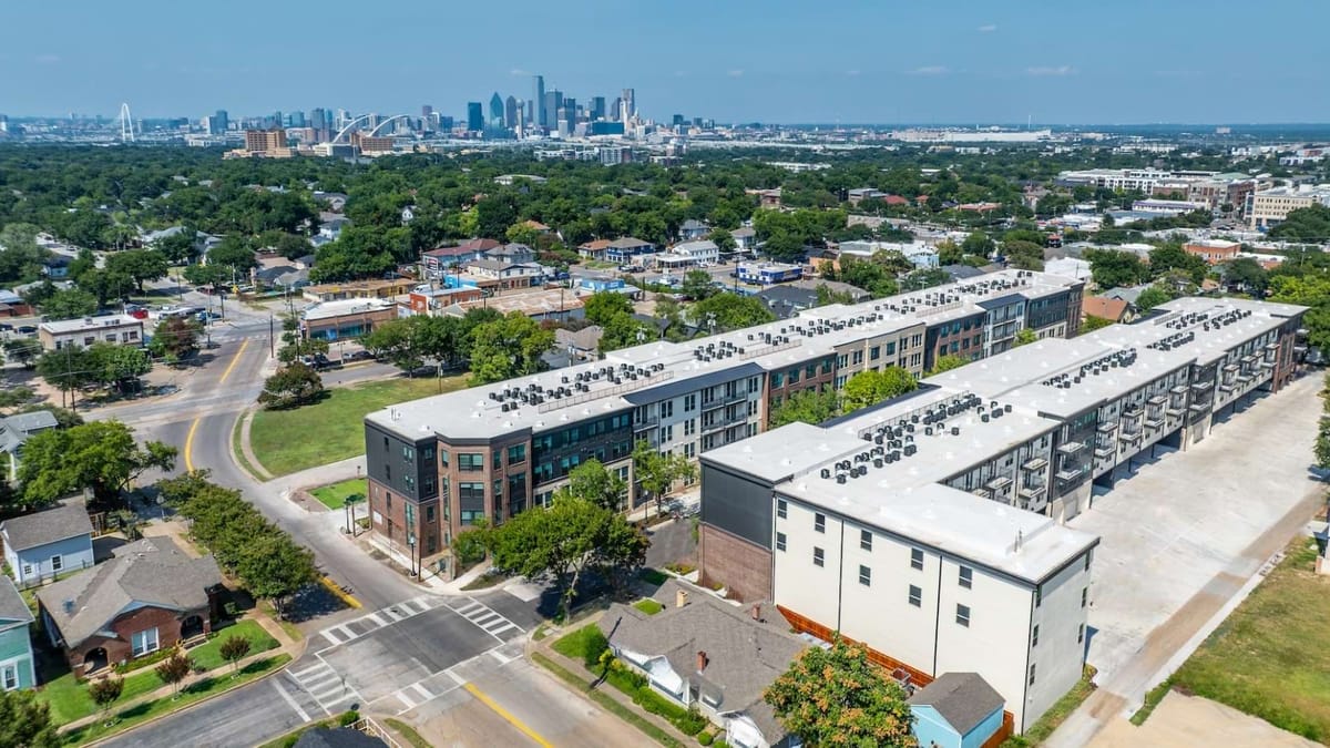 Aerial view of two long four-story apartment buildings among houses and trees, with the downtown Dallas skyline in the distance