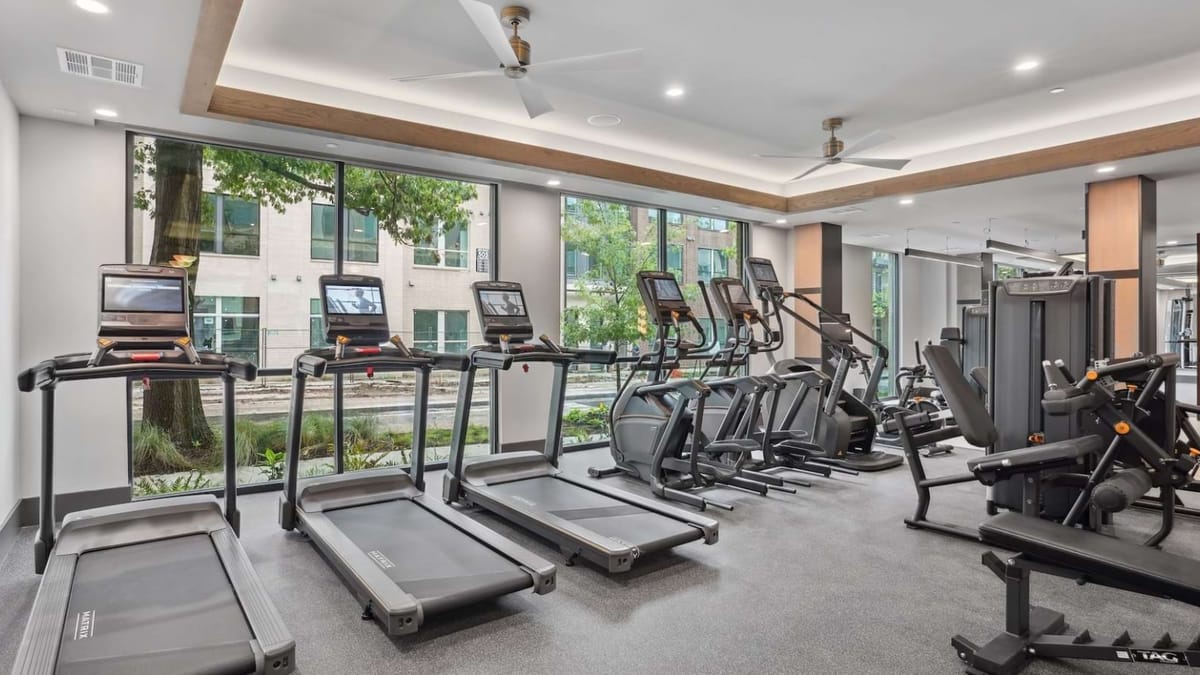 Fitness room with treadmills and elliptical machines facing floor-to-ceiling windows, under a recessed ceiling with fans