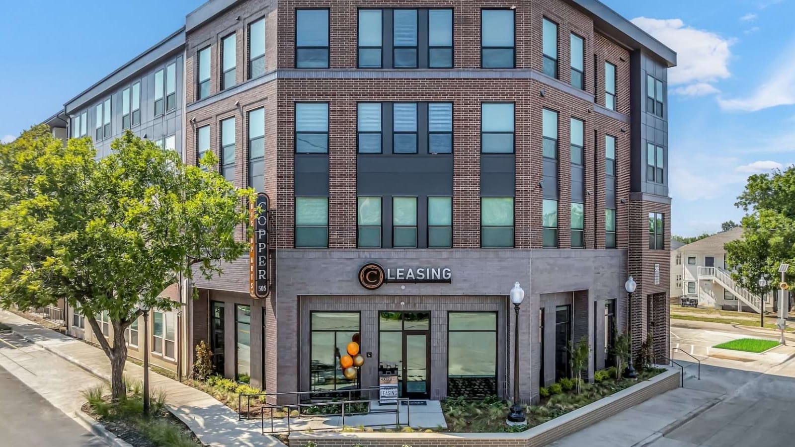 Corner of a four-story brick apartment building with large dark-framed windows and a ground-floor office under a sign reading Leasing, with street trees on the sidewalk