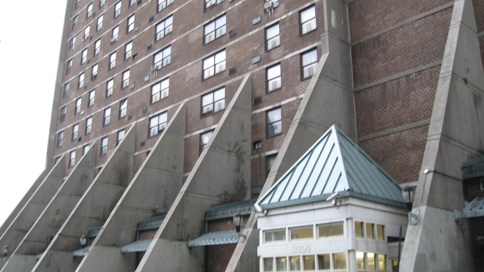 Lower floors of a tall brown-brick apartment tower with rows of windows, tall triangular concrete buttresses along its base, and a small glass entrance pavilion with a green metal pyramid roof marked 3204