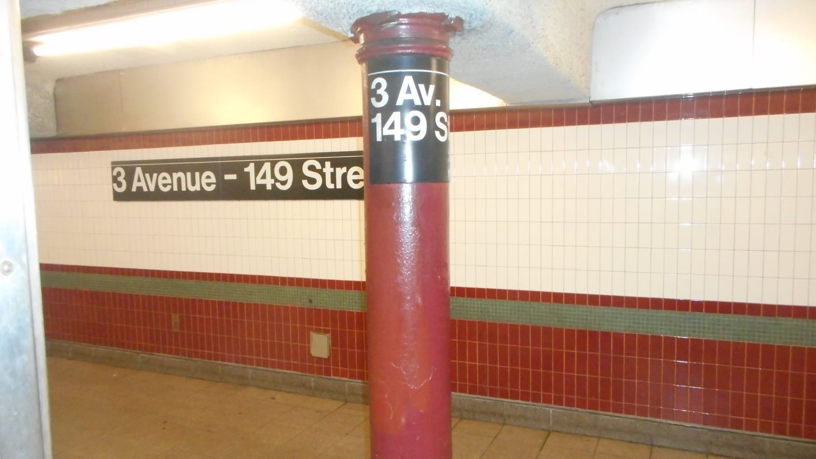 A subway platform with a maroon column labeled 3 Av. 149 St, a black wall sign reading 3 Avenue - 149 Street, white tiles above a maroon tile band and a tan tile floor