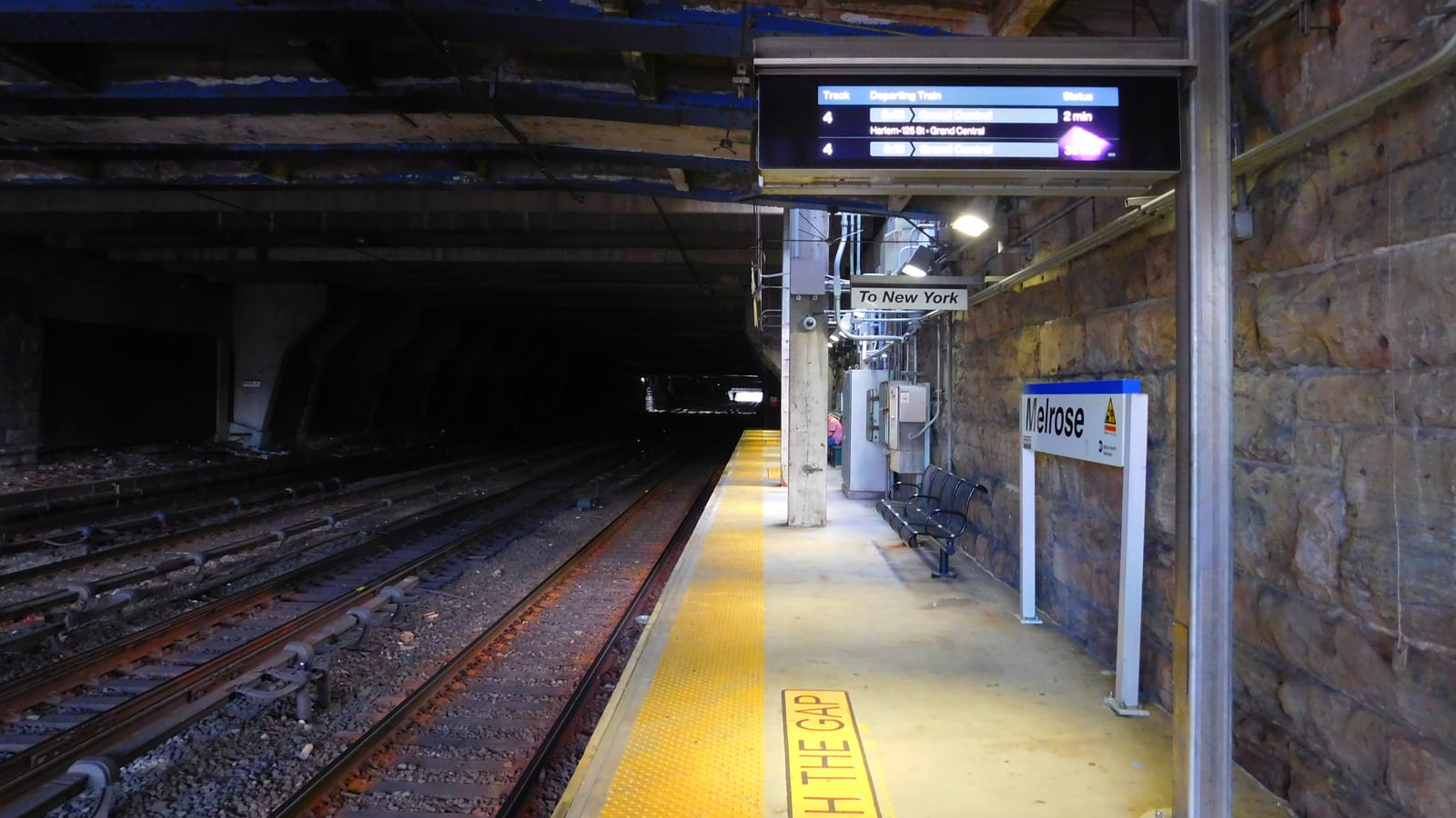 An underground Metro-North platform with a yellow tactile edge strip and the words WATCH THE GAP, a blue-and-white Melrose station sign on a stone wall, a black bench, a departure screen listing trains to Grand Central, and several tracks leading into a dark tunnel
