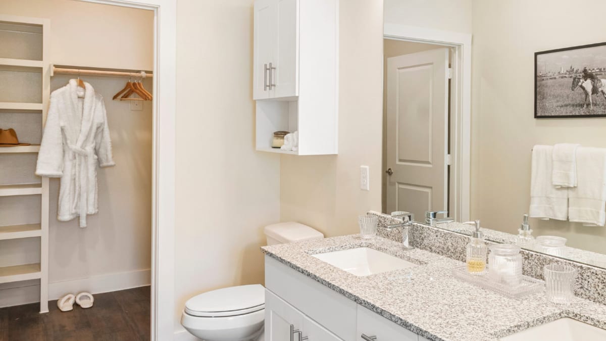 Bathroom with a granite double-sink vanity, white cabinets, a wall cabinet over the toilet and a walk-in closet with built-in shelves