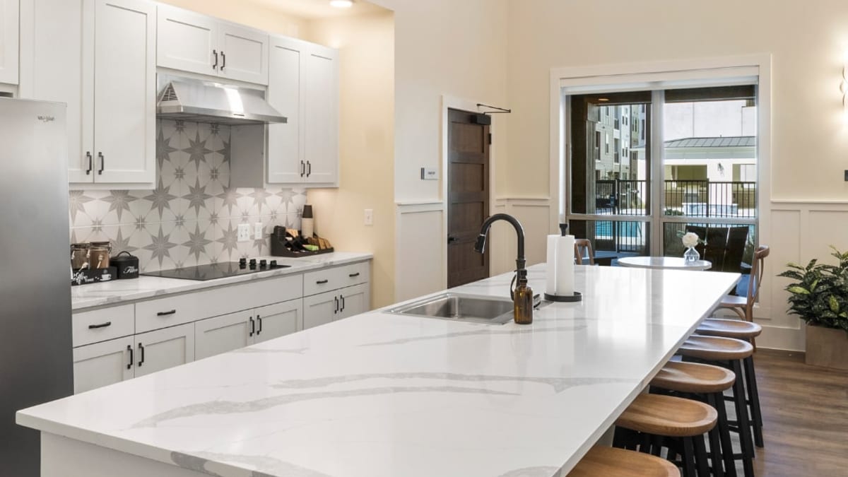Clubhouse kitchen with white cabinets, a patterned tile backsplash, a long island with wood stools and glass doors to a pool courtyard