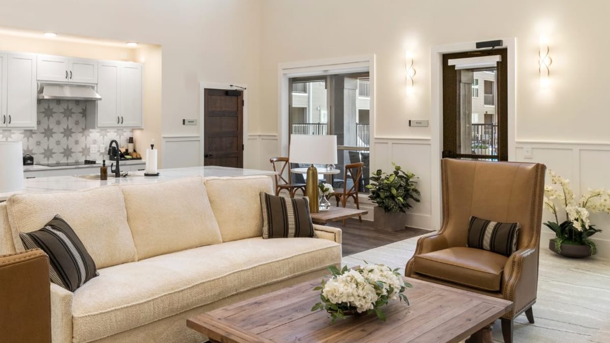 Clubhouse lounge with a cream sofa, a leather armchair, a wood coffee table and a kitchen behind, with doors to a courtyard