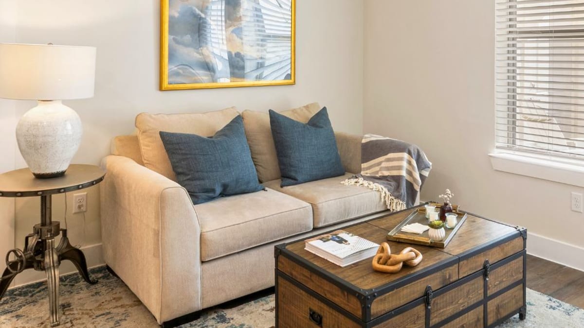 Living room corner with a beige sofa, blue cushions, a trunk used as a coffee table and a window with blinds