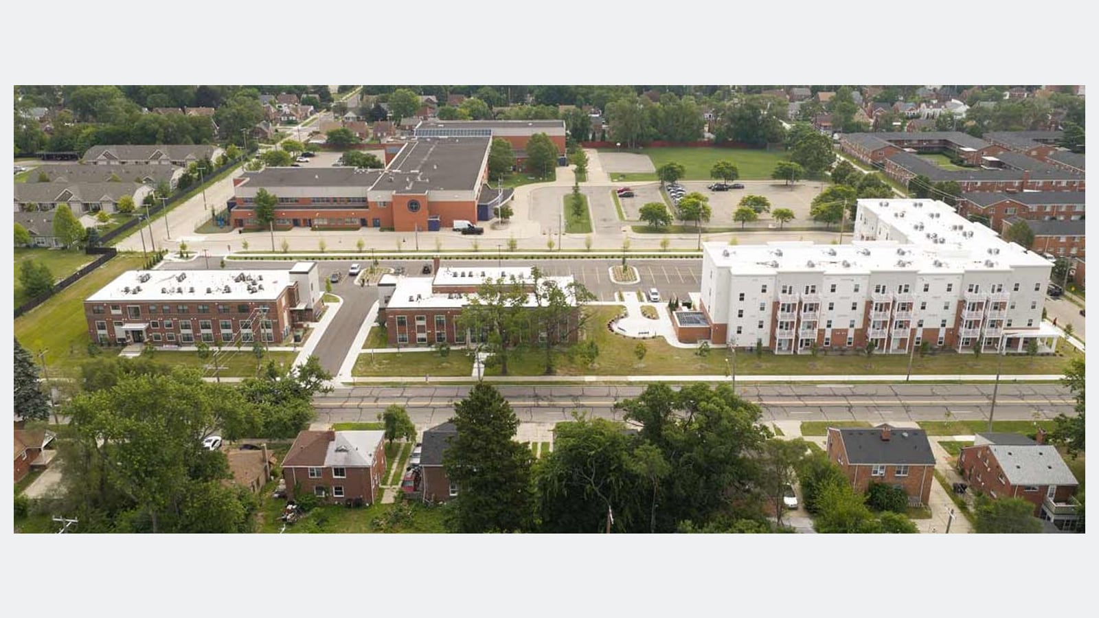 Aerial photograph of a campus with two low red brick buildings on the left and a long four-story white and brick building on the right, with parking between them and houses around