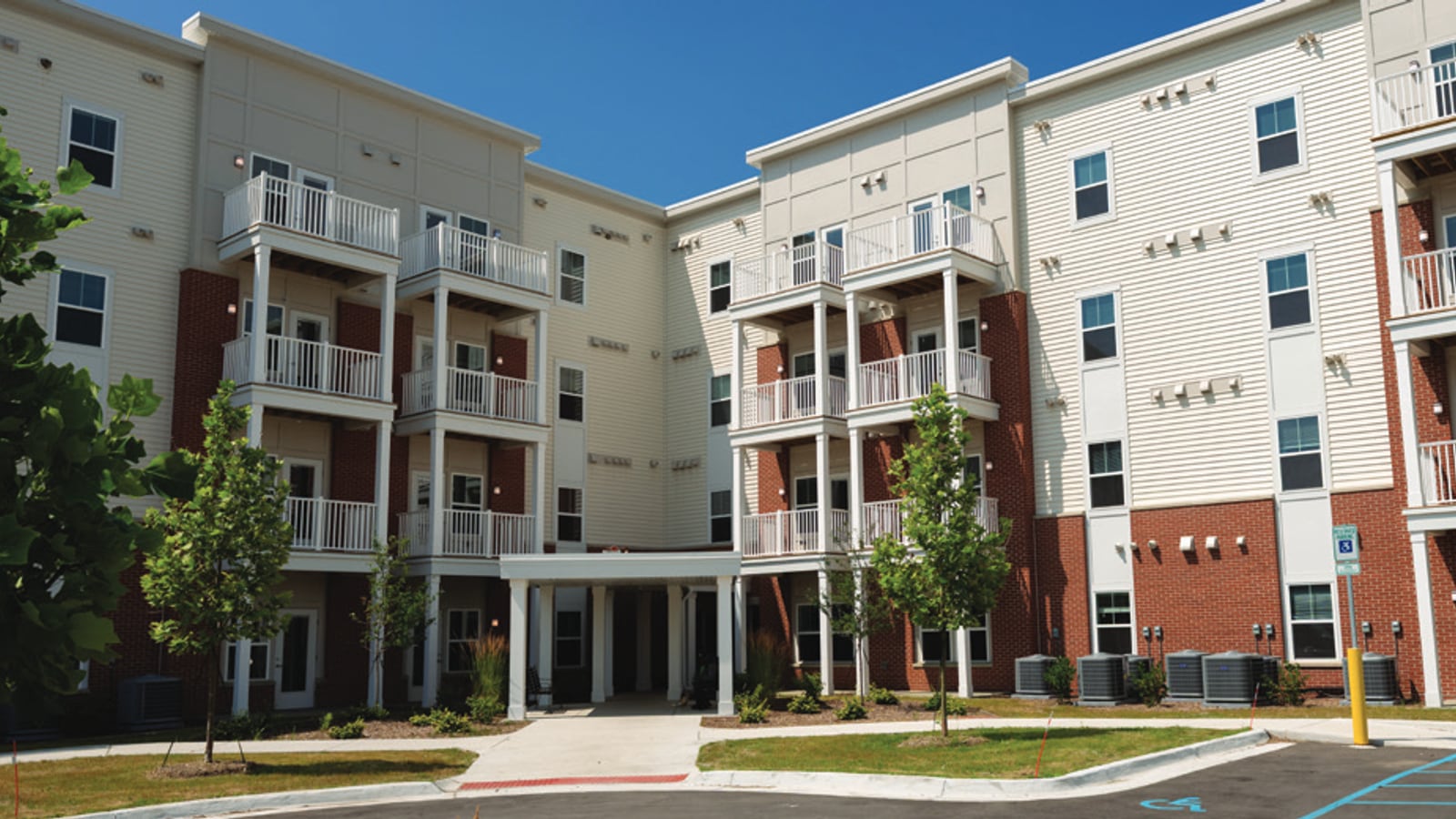 Photograph of a four-story apartment building with white siding, red brick and stacked white balconies around an entrance court, with a parking lot in front