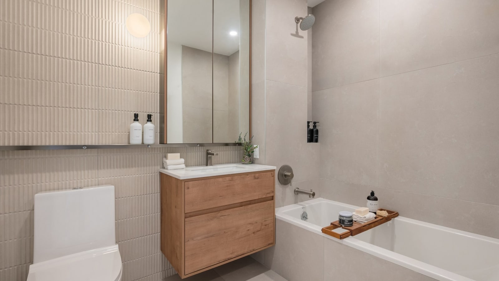 Photograph of a bathroom with ribbed beige wall tile and large grey tiles, a wall-hung wood vanity with a white top, a mirrored cabinet, a white toilet and a bathtub with a wall-mounted shower head and a wooden tray across it