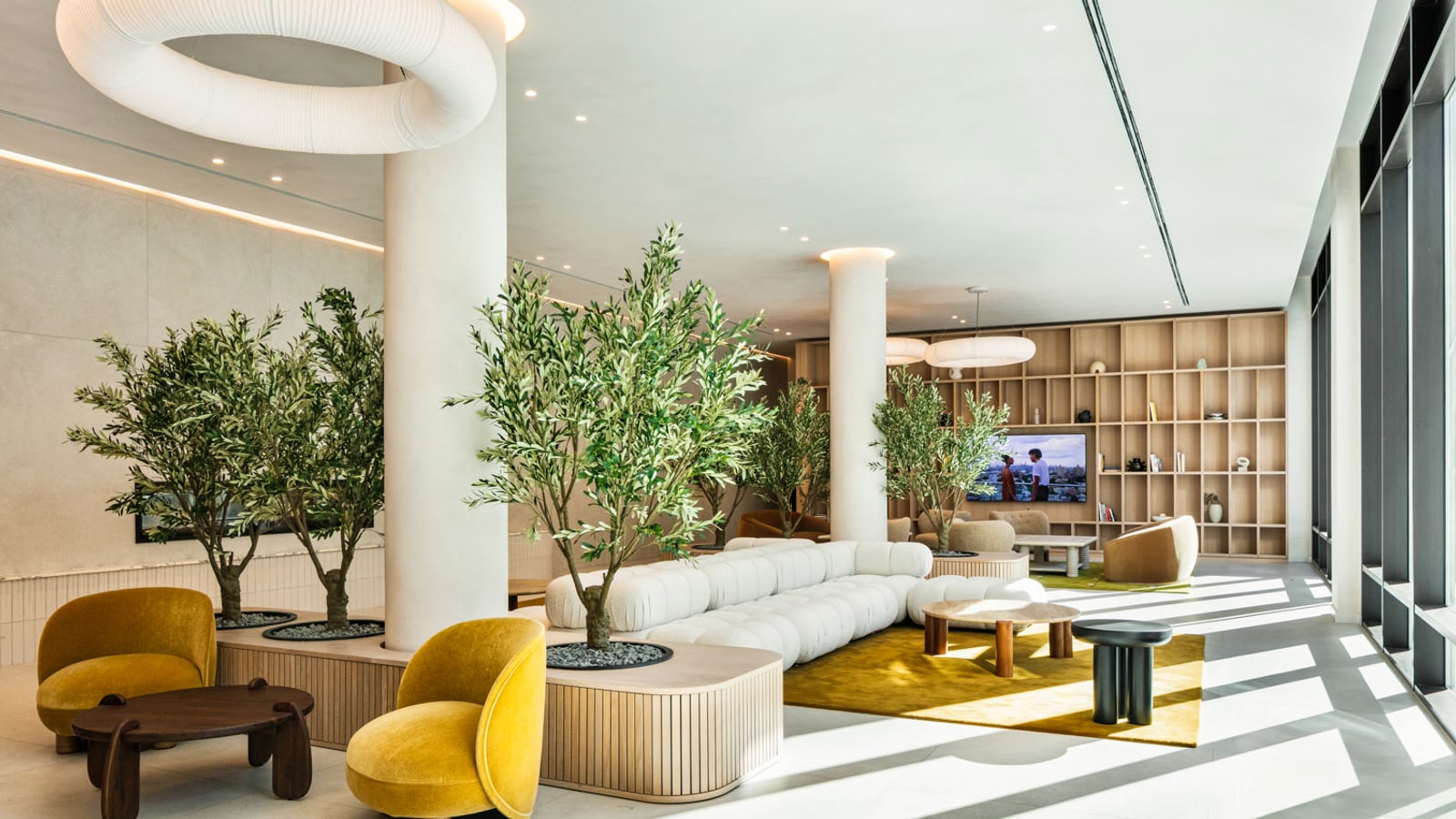 Photograph of a large sunlit lounge with round white columns, a ring-shaped white ceiling light, potted olive trees in pale planters, mustard armchairs, a long white sofa on a yellow rug, a wall of wooden shelves with a television and floor-to-ceiling windows at the right