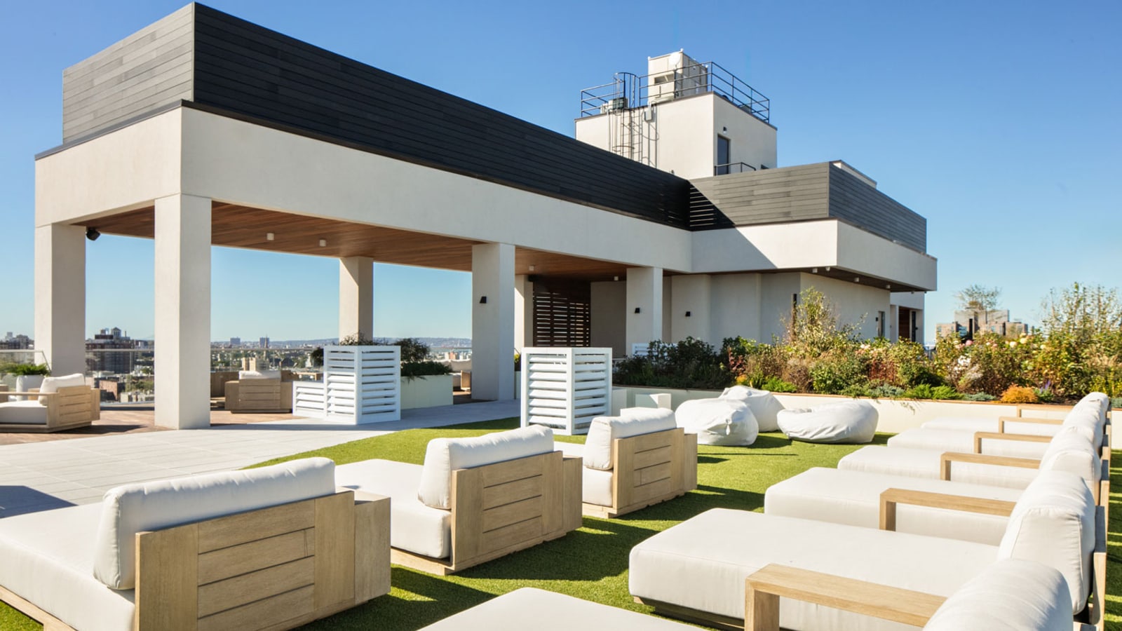 Photograph of a roof terrace under a clear sky with white cushioned lounge seats in wood frames on artificial grass, white beanbag seats, planted beds, a covered area under a white and dark-clad rooftop structure and low buildings on the horizon