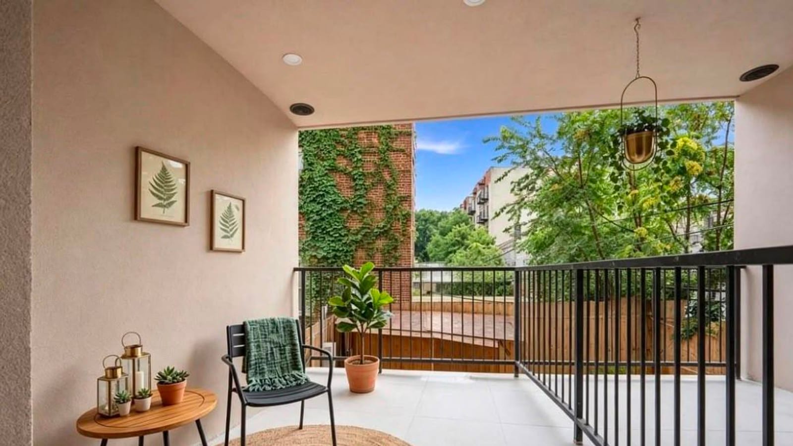 A covered balcony with pale pink walls, a tiled floor and a black metal railing. A small wooden table with lanterns and succulents, a dark chair with a green throw and a potted fig tree stand on a round jute rug. Beyond the railing are trees, an ivy-covered brick wall, a wooden fence and a row of apartment buildings with balconies.