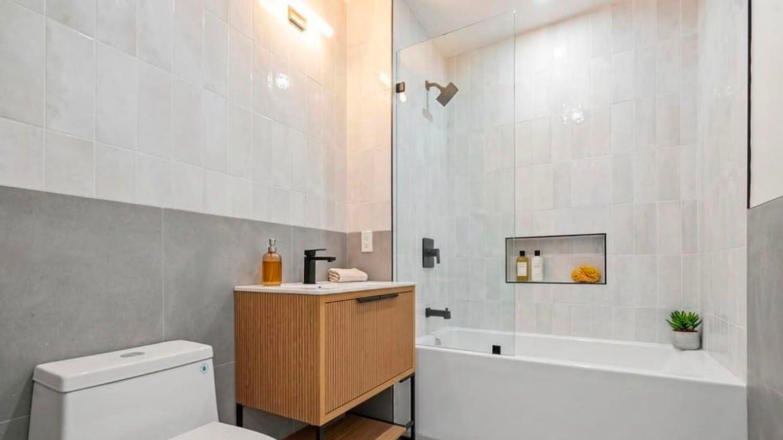 A small bathroom with large grey tiles on the lower walls and white tiles above, a wooden vanity with a fluted front and a black faucet, a white toilet in the foreground, and a bathtub with a glass shower screen, a black shower head and a tiled shelf niche holding bottles.