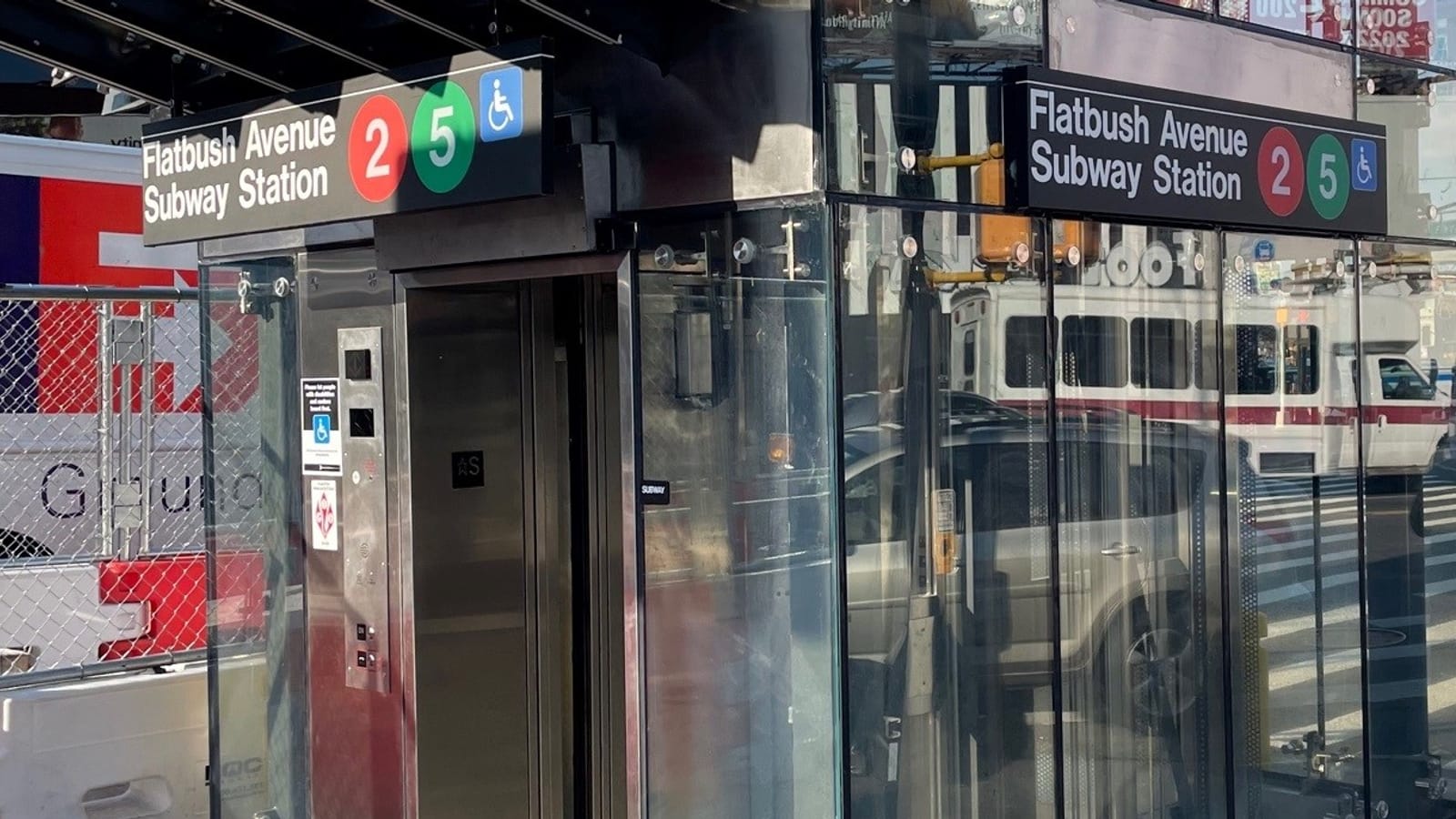 A glass and steel elevator kiosk on a sidewalk with open steel doors, a call panel and signs reading Flatbush Avenue Subway Station with the 2 and 5 line symbols and a wheelchair symbol. Cars and a van are reflected in the glass.