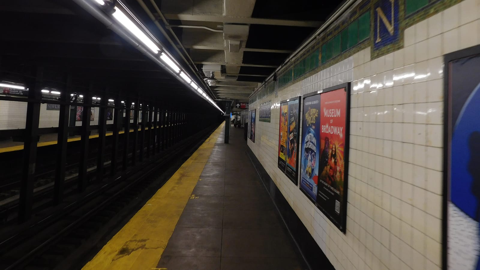 An empty underground subway platform with a yellow edge strip, fluorescent lights along the ceiling and white tiled walls with a green mosaic band and a large letter N. Posters hang on the wall at right, and the platform across the tracks carries a sign reading Newkirk.