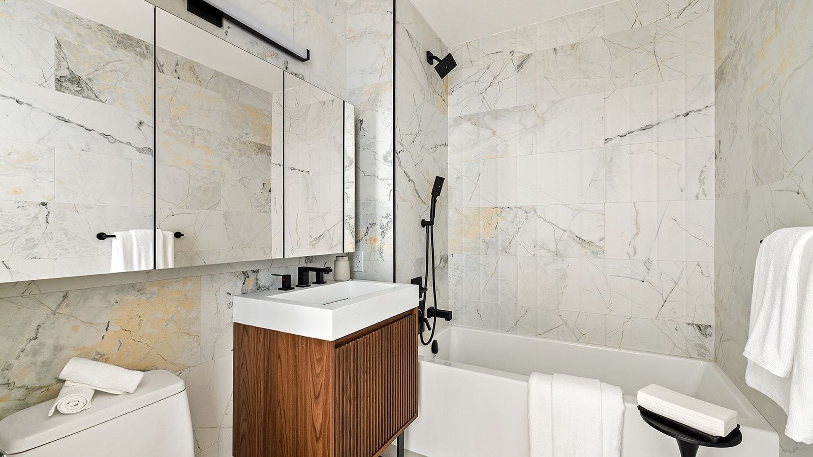 Photograph of a bathroom with marble-look wall tile, a white sink on a ribbed wood vanity, black fixtures, a mirrored cabinet, a white toilet and a white bathtub with a handheld shower