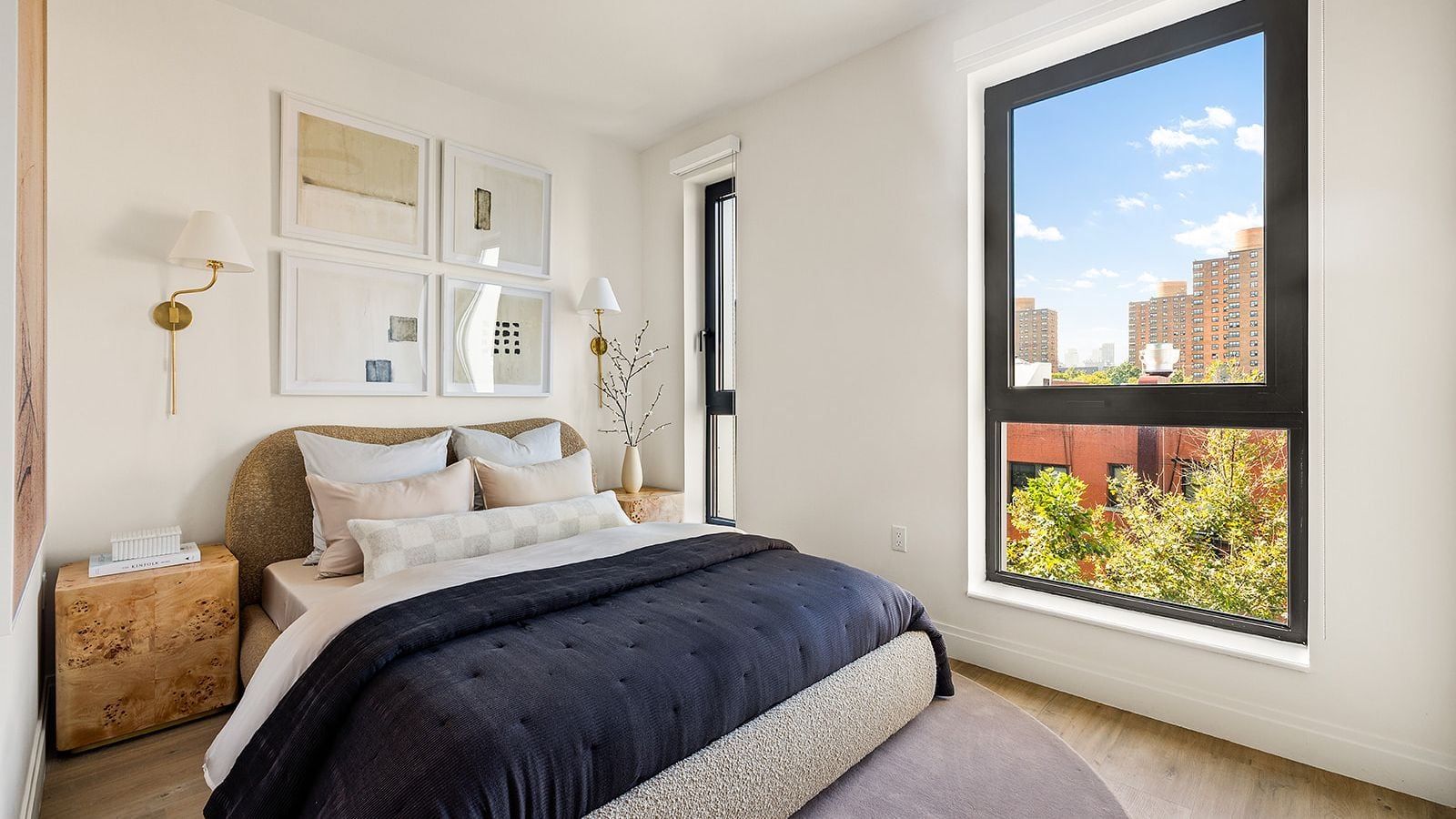 Photograph of a furnished bedroom with a bed in dark blue bedding, wood bedside tables, brass wall lamps, four framed prints and a large black-framed window looking onto brick apartment towers and treetops