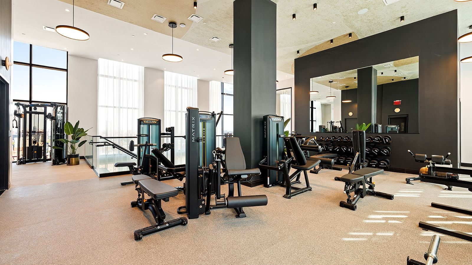 Photograph of a gym with black weight machines and benches, a rack of dumbbells under a wall mirror, a dark square column, round ceiling lights and tall curtained windows