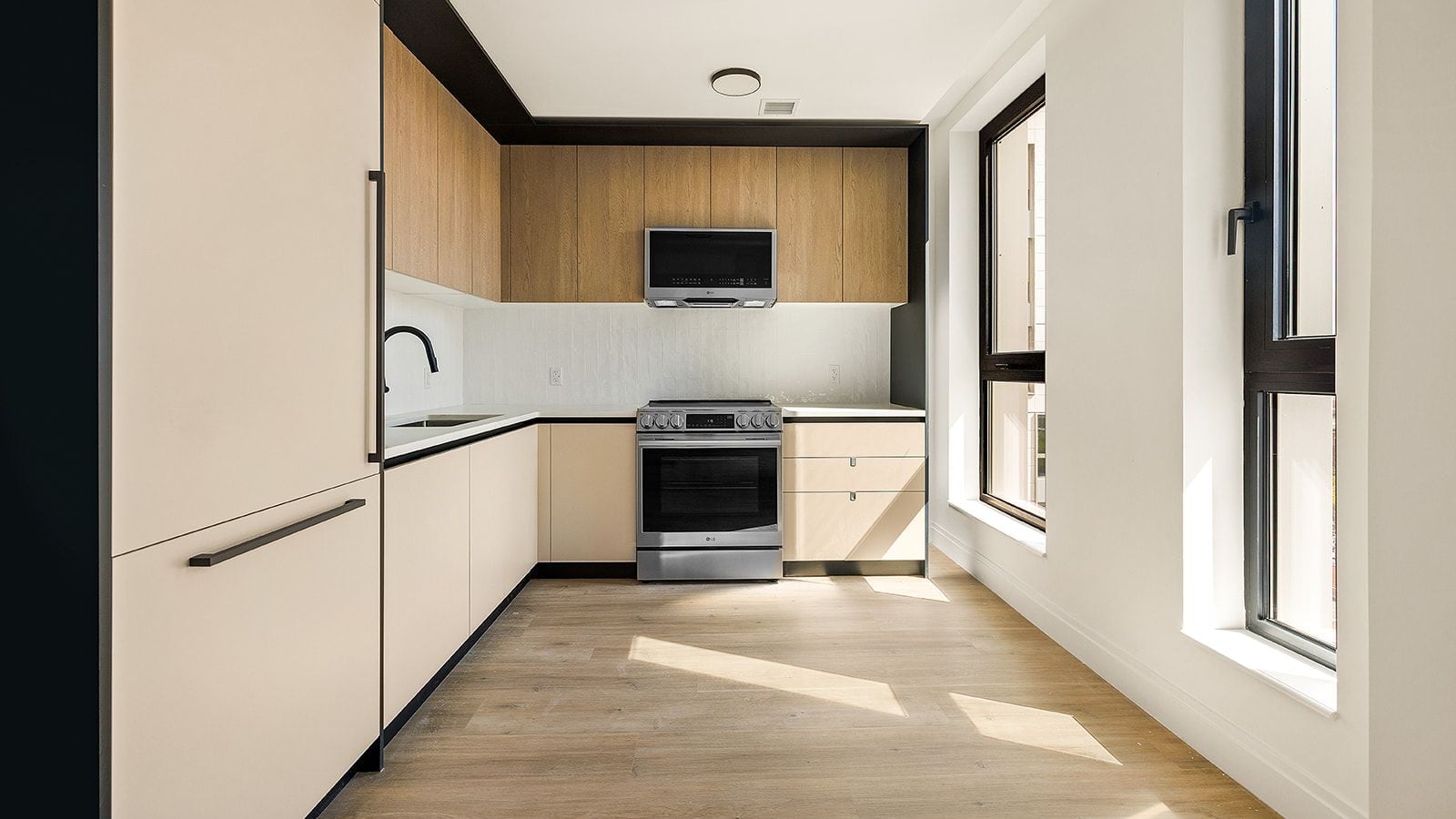 Photograph of an empty kitchen with flat cream lower cabinets, wood-tone upper cabinets, a white tile backsplash, a stainless-steel microwave above a stainless-steel range with front knobs and a flat cooktop, a panelled refrigerator at the left and two tall windows at the right