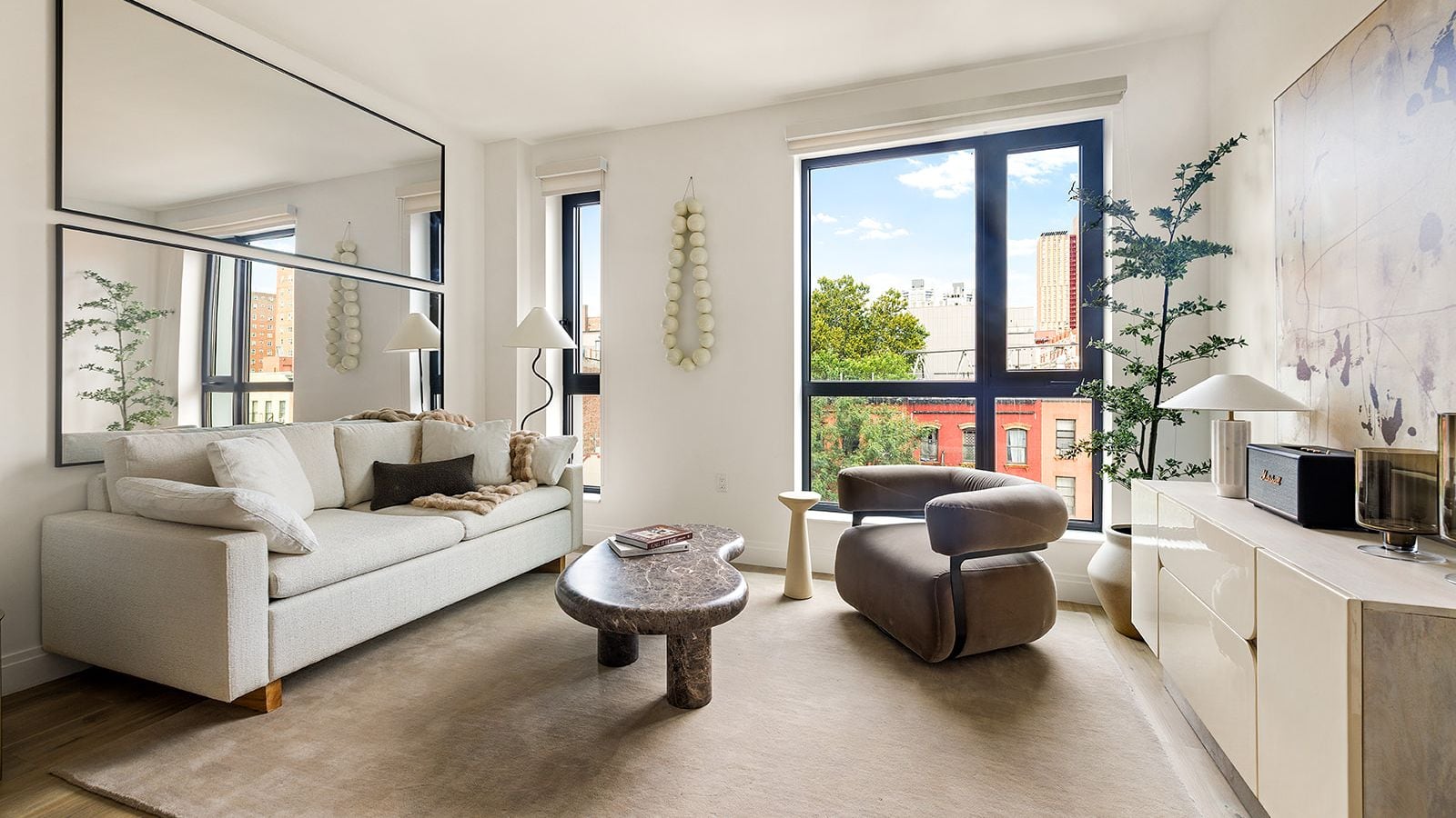 Photograph of a furnished living room with a white sofa under two long wall mirrors, a stone coffee table, a brown armchair, a pale sideboard and a tall black-framed window looking onto red brick buildings and trees