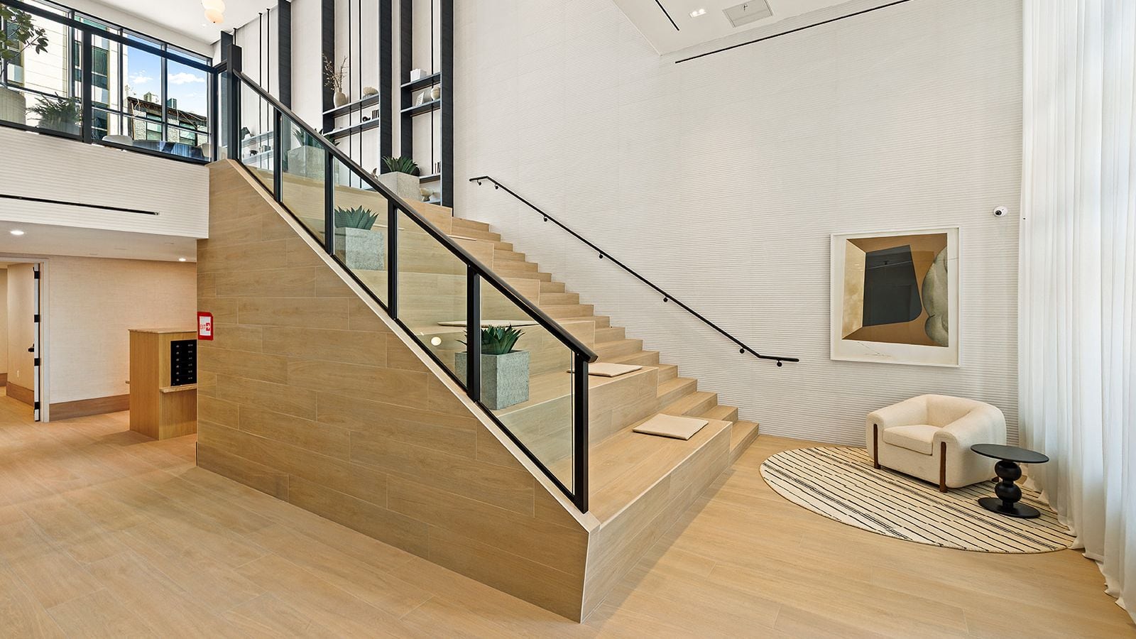 Photograph of a double-height lobby with a wide wood-clad stair of seating steps behind a glass railing, planters and cushions on the steps, a framed artwork on a white ribbed wall, a cream armchair on a striped round rug and a mezzanine with black-framed windows above