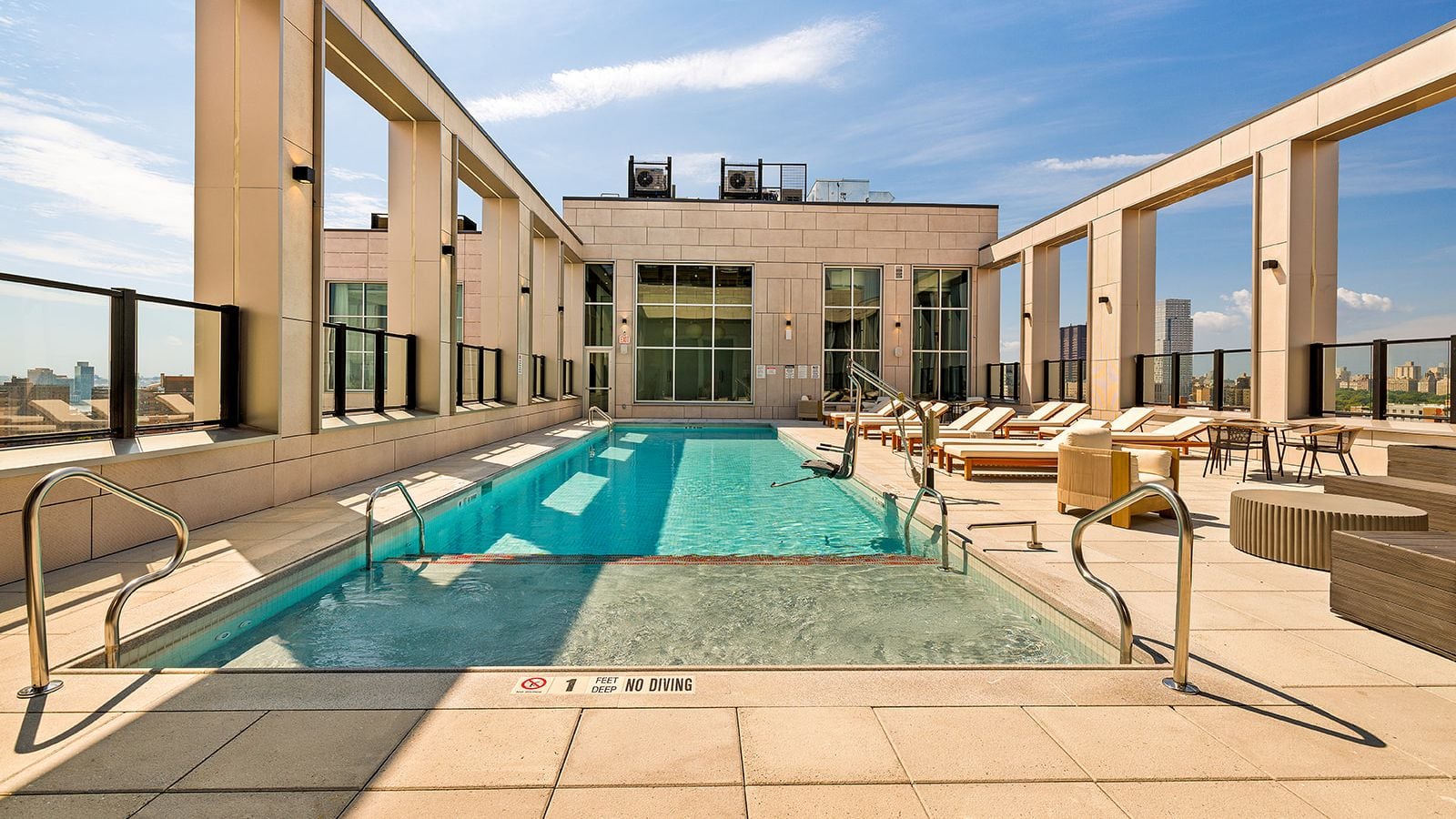 Photograph of a rooftop swimming pool with steel handrails and a pool lift, lounge chairs along the right side, a glass-fronted rooftop room at the far end, tan stone columns framing the sky and city buildings in the distance; a marker on the pool edge reads 1 feet deep, no diving
