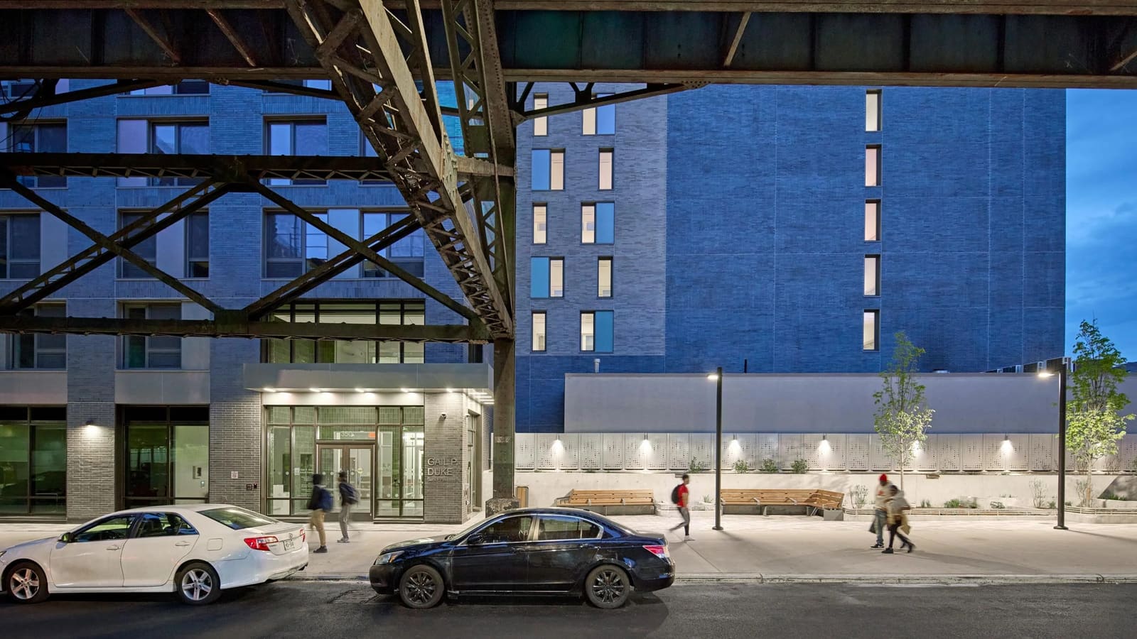 Photograph at dusk taken from under steel subway tracks, showing the grey brick base of an apartment building with a lit glass entrance, metal letters reading Gail P. Duke Senior Residence beside the doors, wooden benches and a planter along the sidewalk at the right, two parked cars and people walking