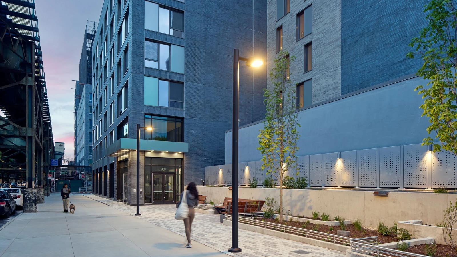 Photograph at dusk of a wide sidewalk beside a dark grey brick apartment building with a lit entrance under a canopy, the number 350 over the doors, street lamps, young trees, wooden benches and planters along a concrete wall, subway columns at the left and a person walking a dog in the distance