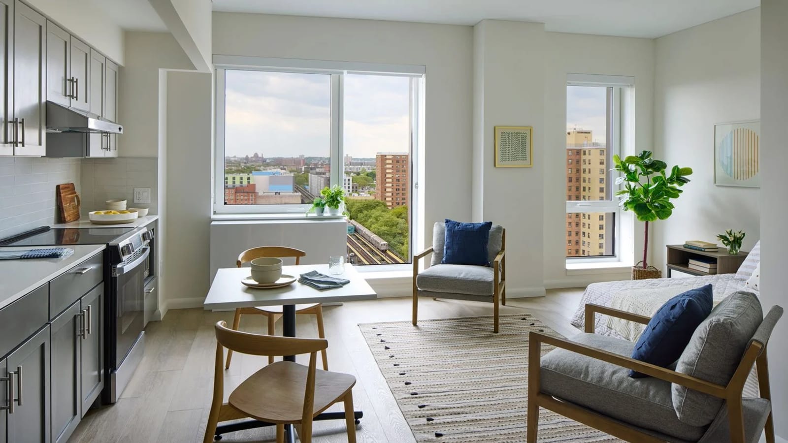 Photograph of a furnished apartment with grey kitchen cabinets, a smooth-top electric range and a range hood along the left wall, a small table with two wooden chairs, two armchairs, a bed at the right and large windows looking out over subway tracks and brick apartment towers