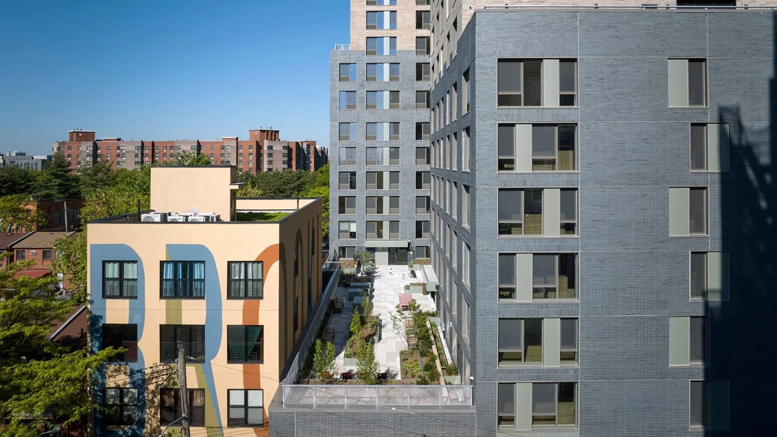 Photograph of a long planted terrace with paving, young trees and seating set between grey brick wings of an apartment building, beside a four-story yellow building with a large painted mural, with brick apartment towers in the distance