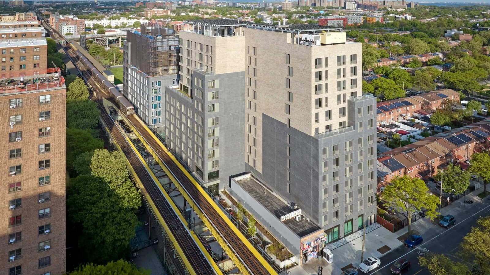 Aerial photograph of a 13-story apartment building with pale beige brick upper floors and grey brick lower floors and solar panels on its roofs, beside two raised subway tracks with yellow edges at the left, a one-story corner store with a Deli & Market sign at its base, rows of two-story houses with solar panels at the right and tall brick apartment towers in the distance