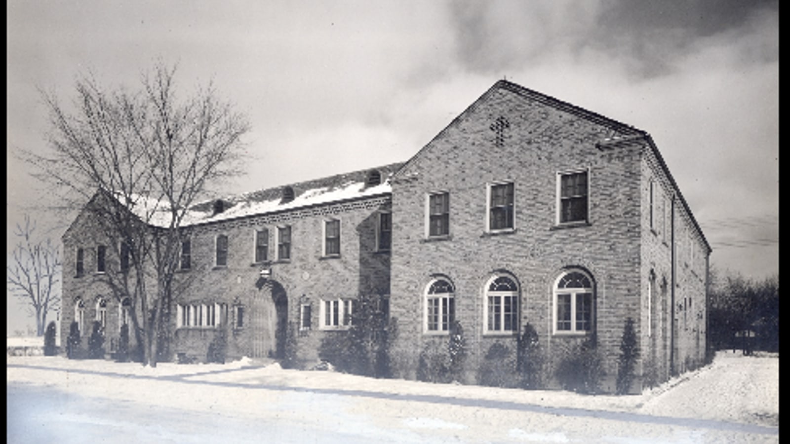 Black-and-white photograph of a two-story brick building with a gabled end, arched ground-floor windows and bare trees, in snow