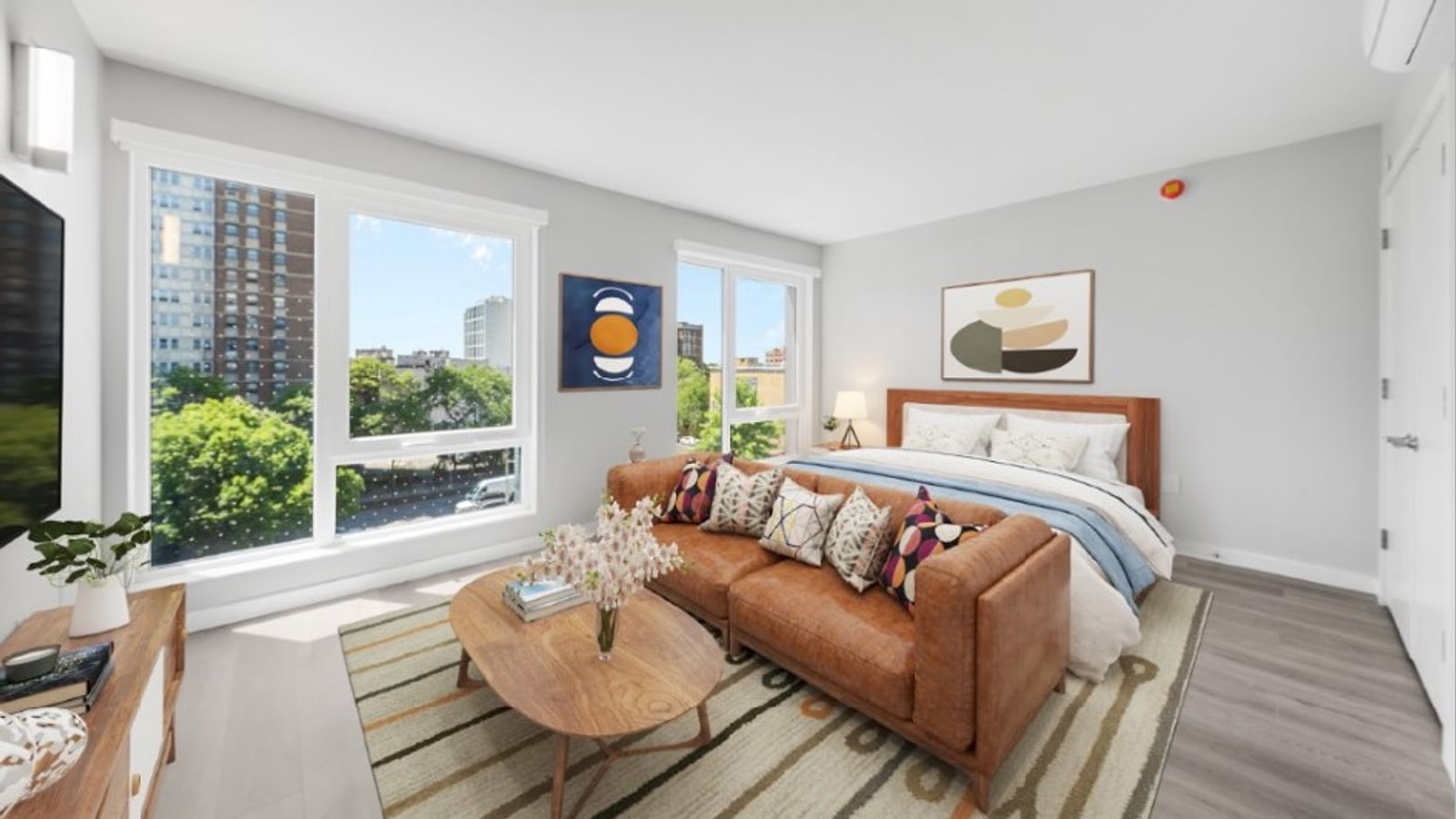 Virtual rendering of a studio apartment with grey plank flooring, two large windows looking over treetops toward apartment towers, a wall-mounted air-conditioning unit, a bed, a tan leather sofa and a round wooden coffee table