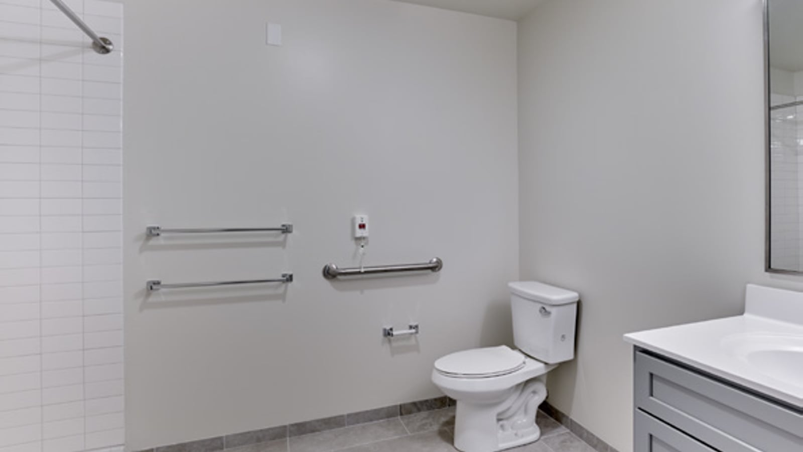 Bathroom with grab bars on the wall, a toilet, a gray vanity with a white top, a wall mirror and an open tiled shower