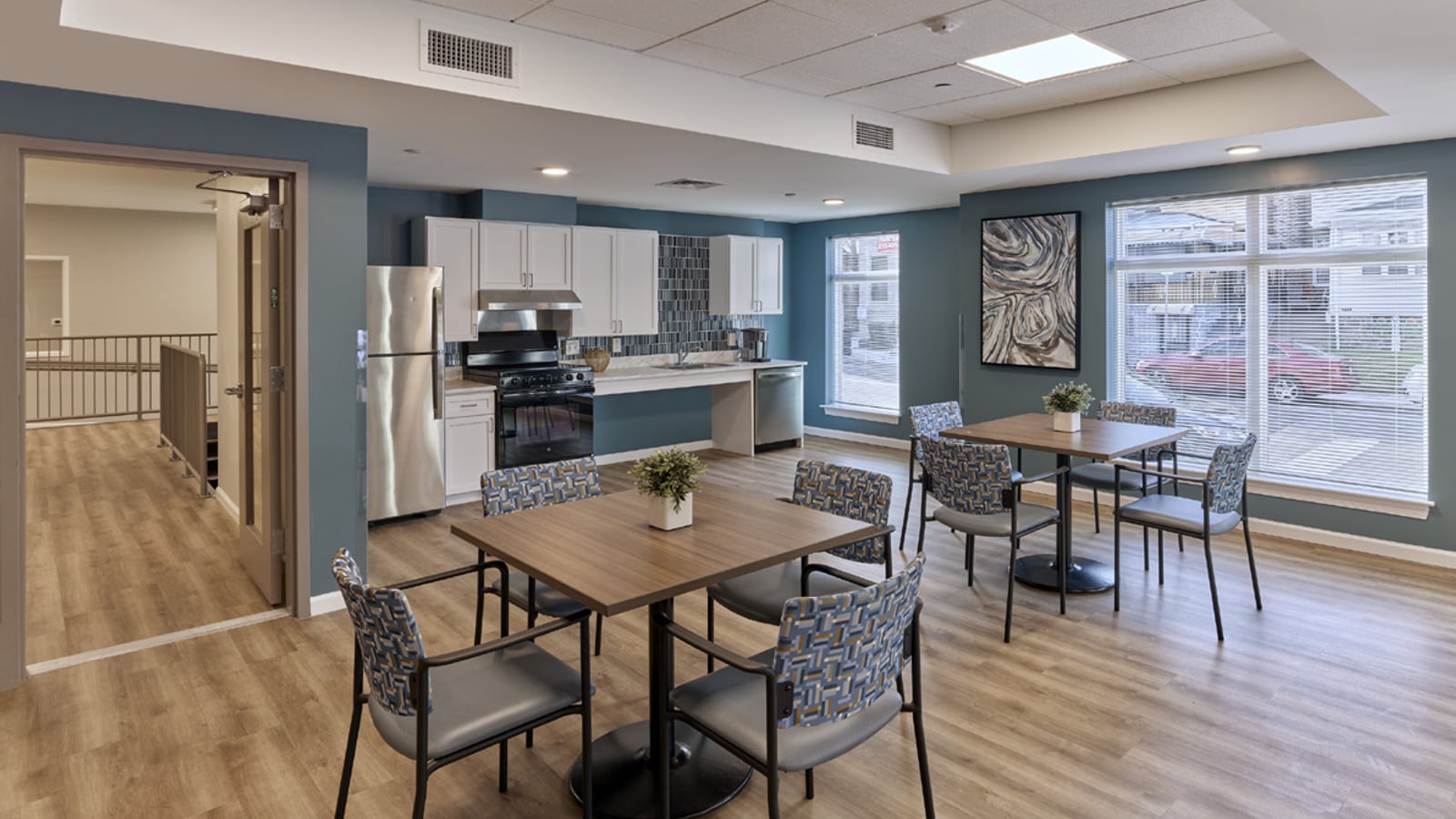 Community room with blue walls, wood-look flooring, two square tables with patterned chairs, a kitchenette with steel appliances and large windows onto the street