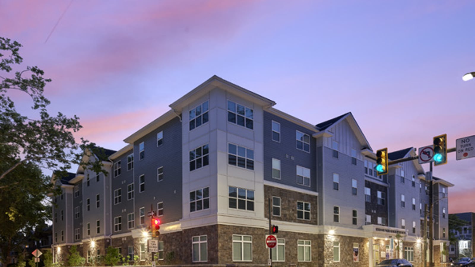 Four-story apartment building with gray siding and a stone ground floor at an intersection with traffic signals, lit at dusk under a pink and purple sky
