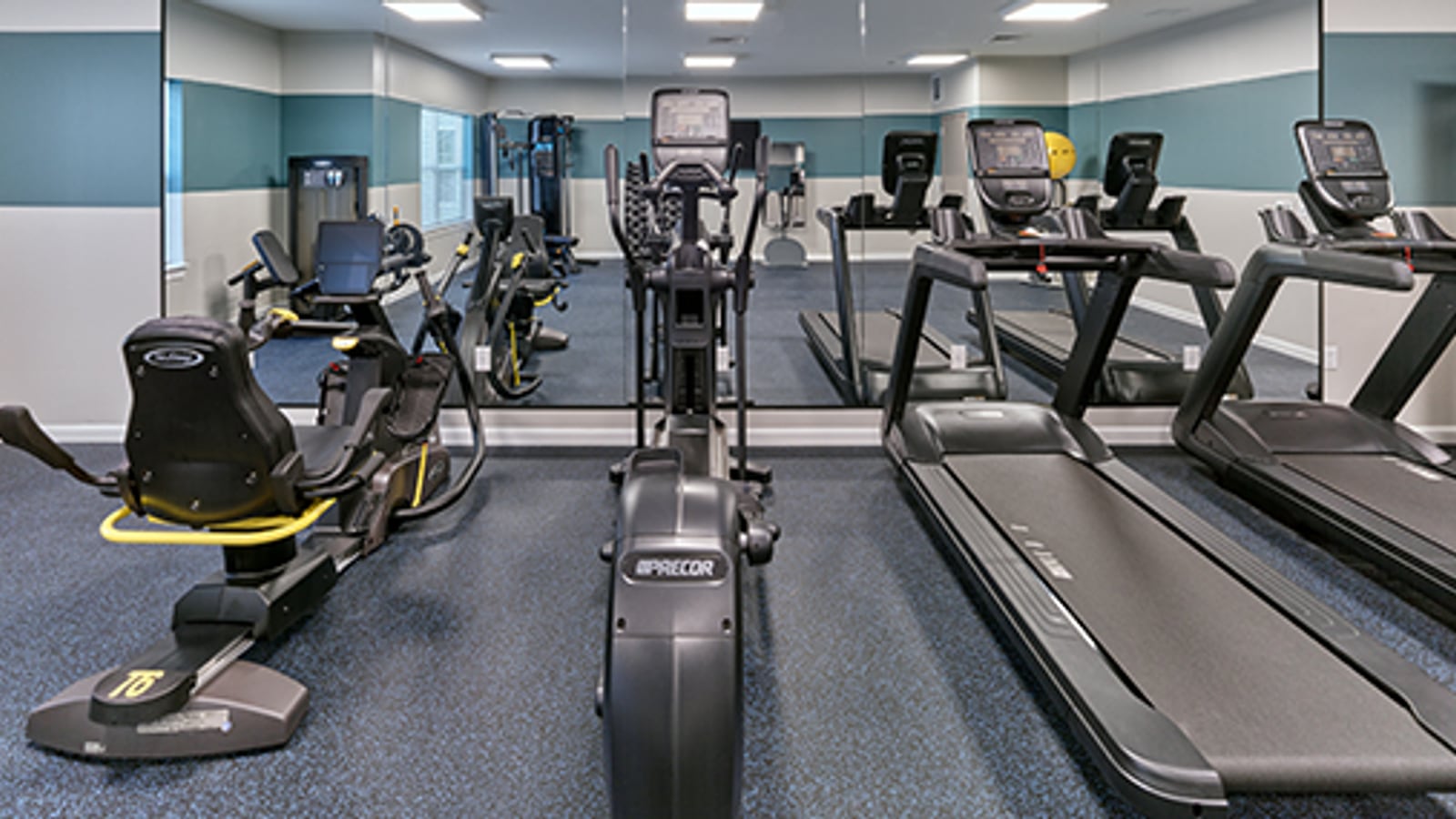 Fitness room with treadmills, an elliptical machine and a recumbent bike on blue flooring, facing a wall of mirrors