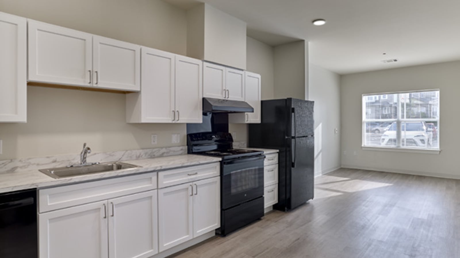 Empty apartment with white kitchen cabinets, a black range, range hood and refrigerator, a steel sink, gray plank flooring and a window at the far end
