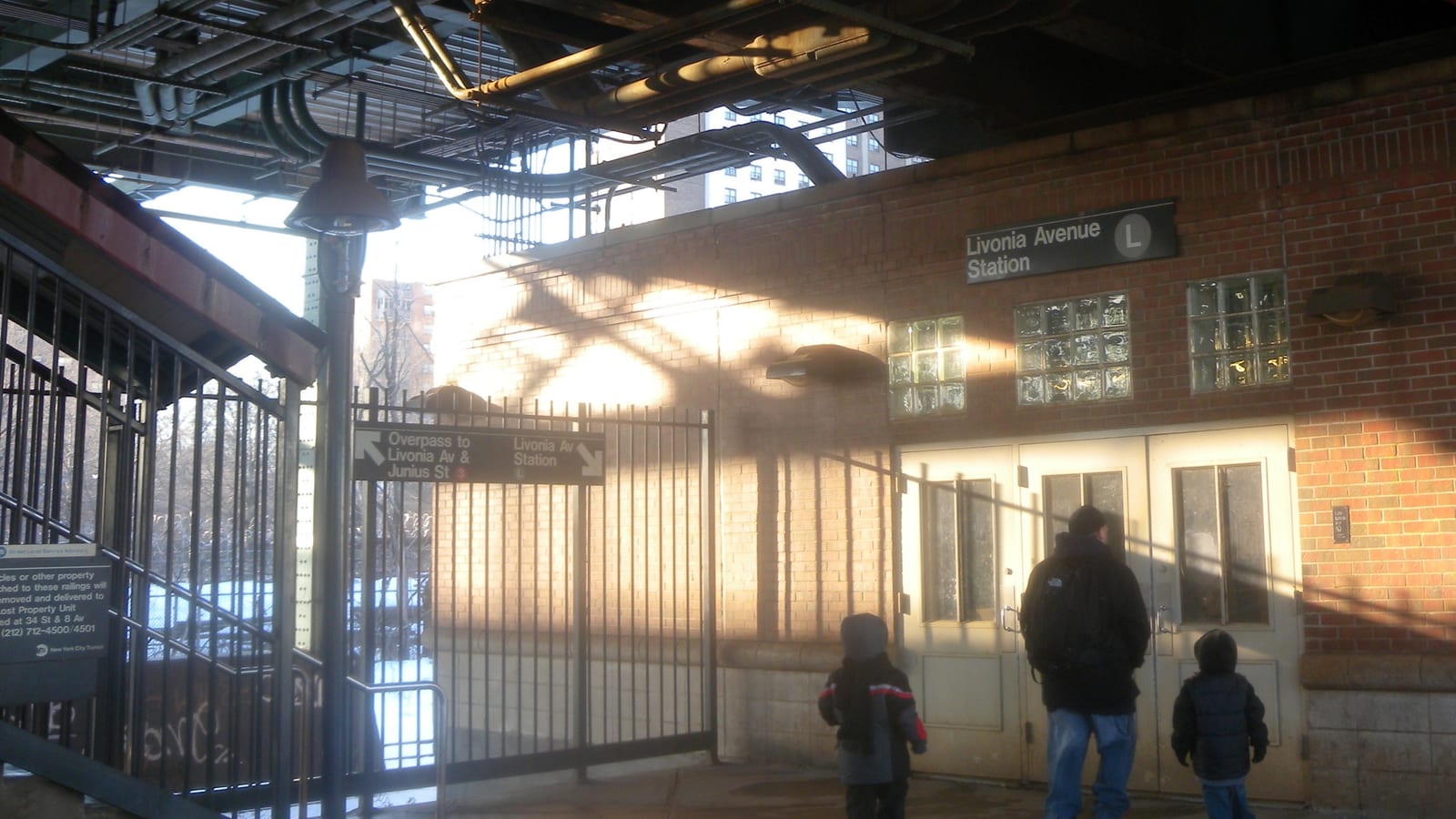 Entrance to the Livonia Avenue L station under the elevated structure, a red brick wall with glass-block windows and a sign reading Livonia Avenue Station L, a sign pointing to the overpass to Livonia Av and Junius St, and an adult and two children seen from behind walking to the doors