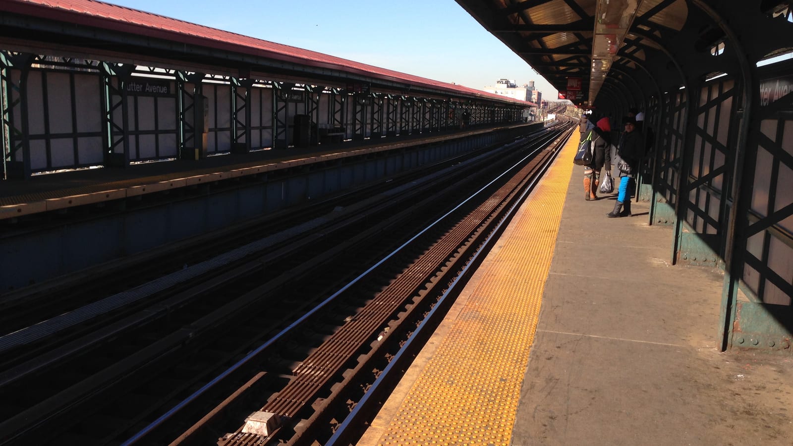 Elevated subway platform under a clear sky, with yellow tactile edging, tracks curving into the distance and a covered platform across the tracks marked Sutter Avenue; a few people wait under the canopy at right