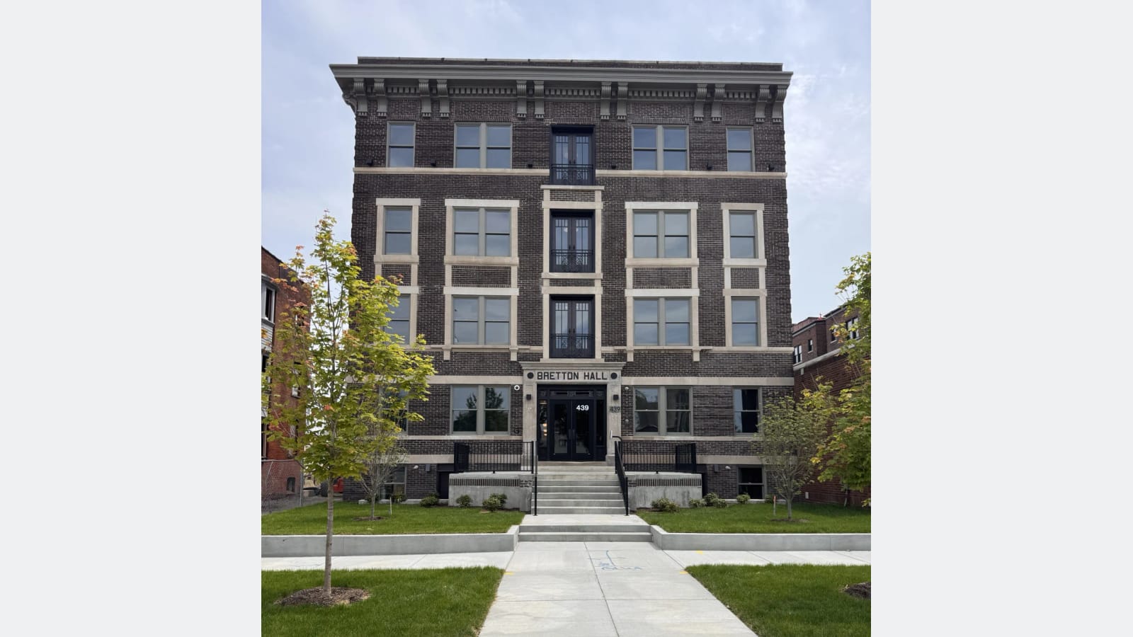 Photograph of a four-story dark brick apartment building with pale stone trim over a raised basement, with the name Bretton Hall and the number 439 above a glass front door, steps, a new sidewalk and young trees