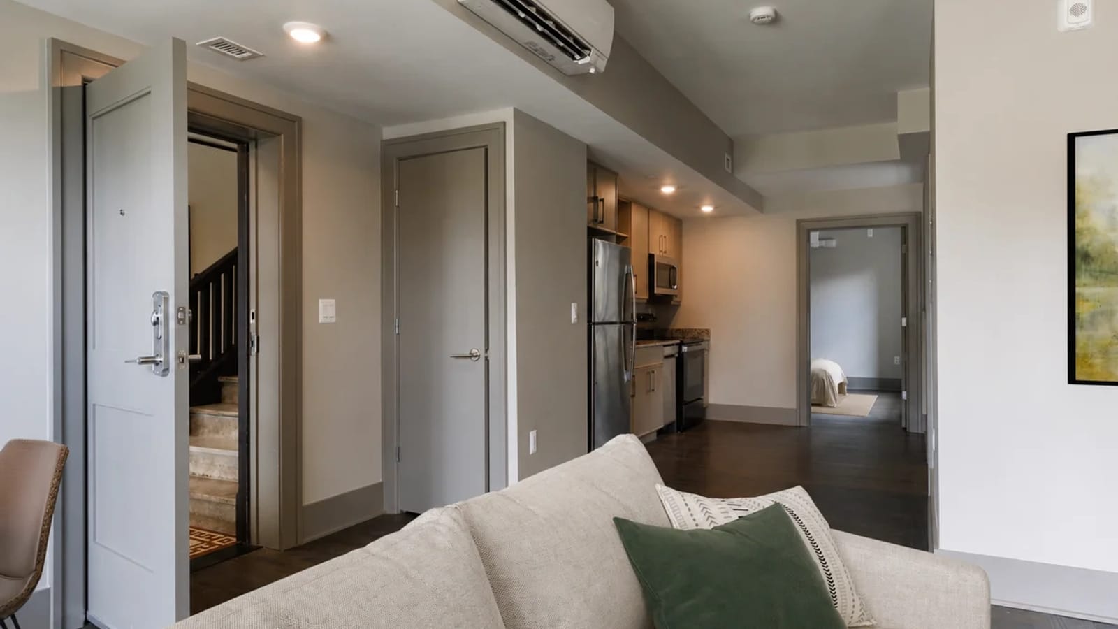 View across a sofa to an apartment's open front door with a staircase beyond it, a closet door, a short kitchen with a stainless steel refrigerator and a doorway to a bedroom, with a wall-mounted air unit near the ceiling