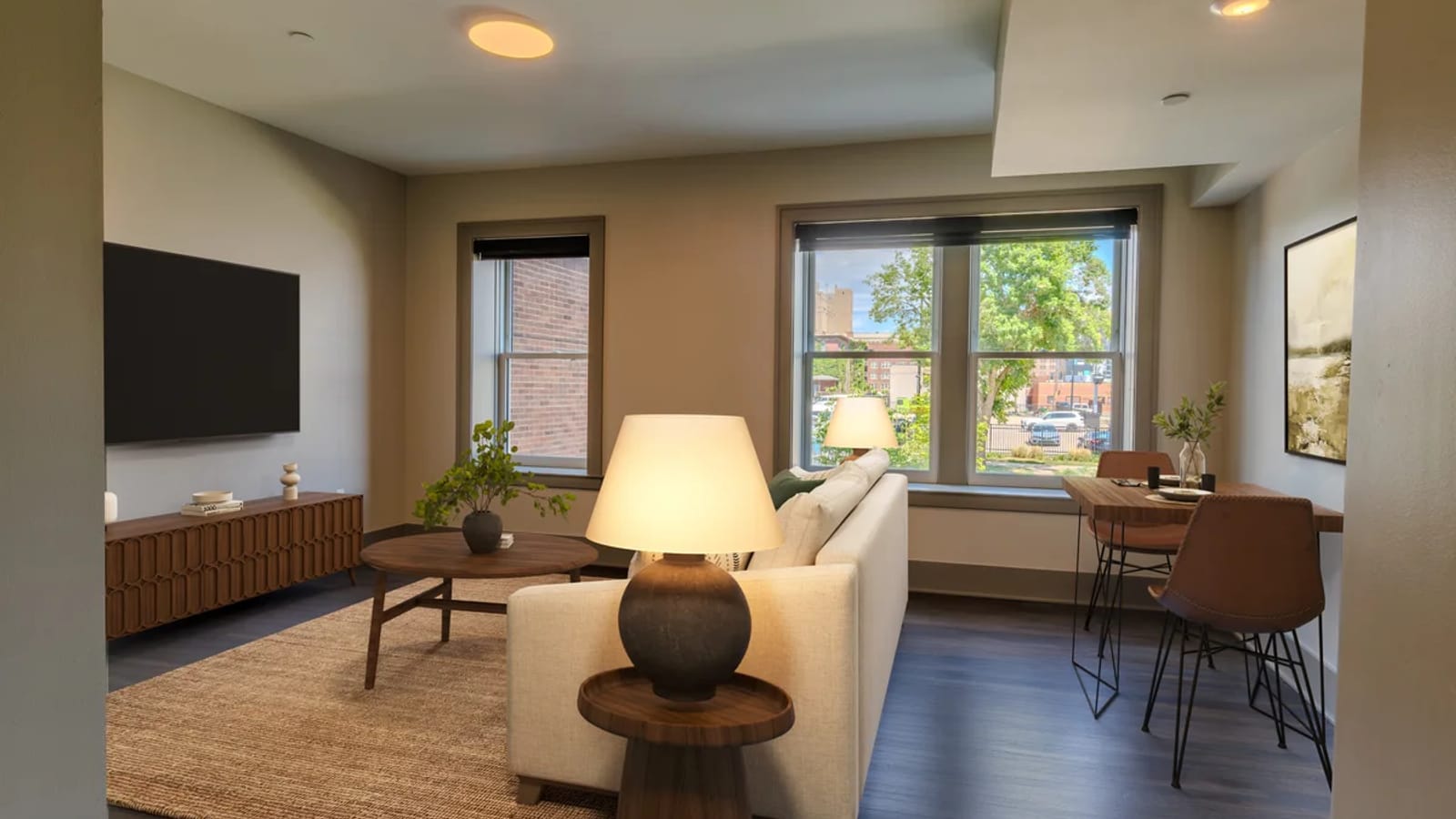 A furnished living room with two tall windows, a wall-mounted television, a sofa, a round coffee table, a lamp and a small dining table with two chairs, on a dark plank floor