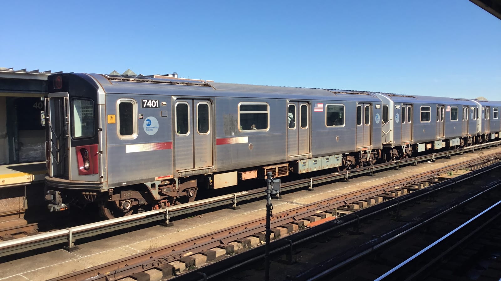 Photograph of a silver 7 train with a red stripe and car number 7401 stopped at an elevated station platform under a clear blue sky, seen across the tracks, with part of a station sign reading 40 at the left