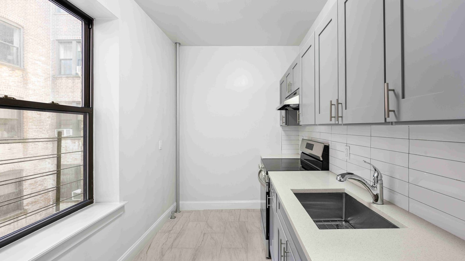 Photograph of an empty galley kitchen with gray cabinets, a white quartz-look counter with a deep steel sink, long white wall tiles, a smooth-top electric range at the far end, a tile floor and a large window on the left looking onto a brick building