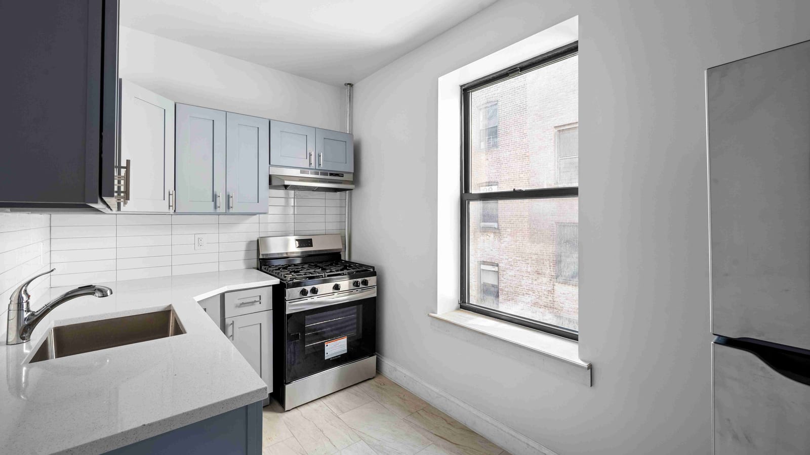 Photograph of a small empty kitchen with gray cabinets, an L-shaped white counter with a sink, a stainless steel gas range with four burners under a range hood, a window looking onto a brick wall, and the edge of a steel refrigerator on the right