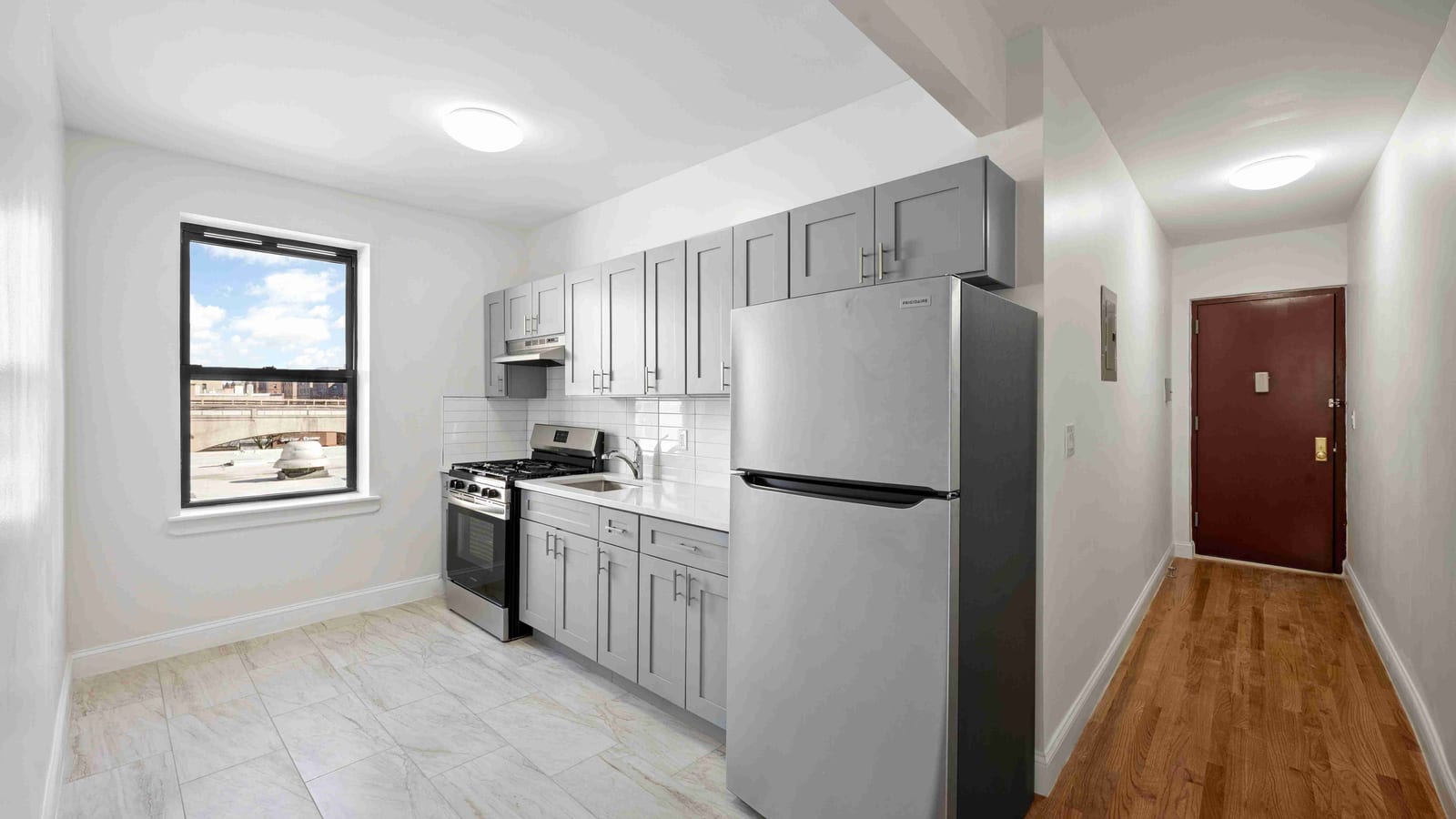 Photograph of an empty kitchen with gray shaker cabinets, a white tile backsplash, a stainless steel gas range, a sink and a tall stainless steel refrigerator, a window on the left showing a rooftop and a blue sky, and a wood-floored hallway on the right leading to a dark red apartment door