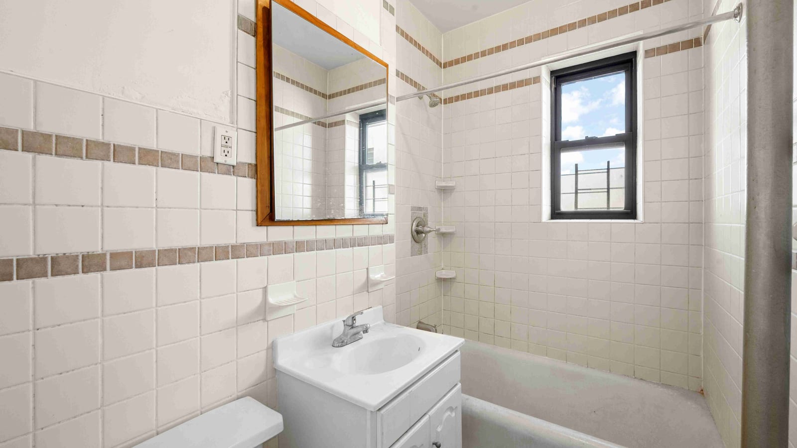 Photograph of a small bathroom with older white square wall tile and a brown accent stripe, a wood-framed mirror, a white vanity sink, and a bathtub with a shower under a small window